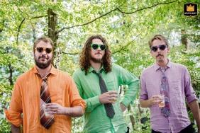 Three male guests in colorful shirts and assorted sunglasses, holding drinks under the trees in Montbonnot, France.